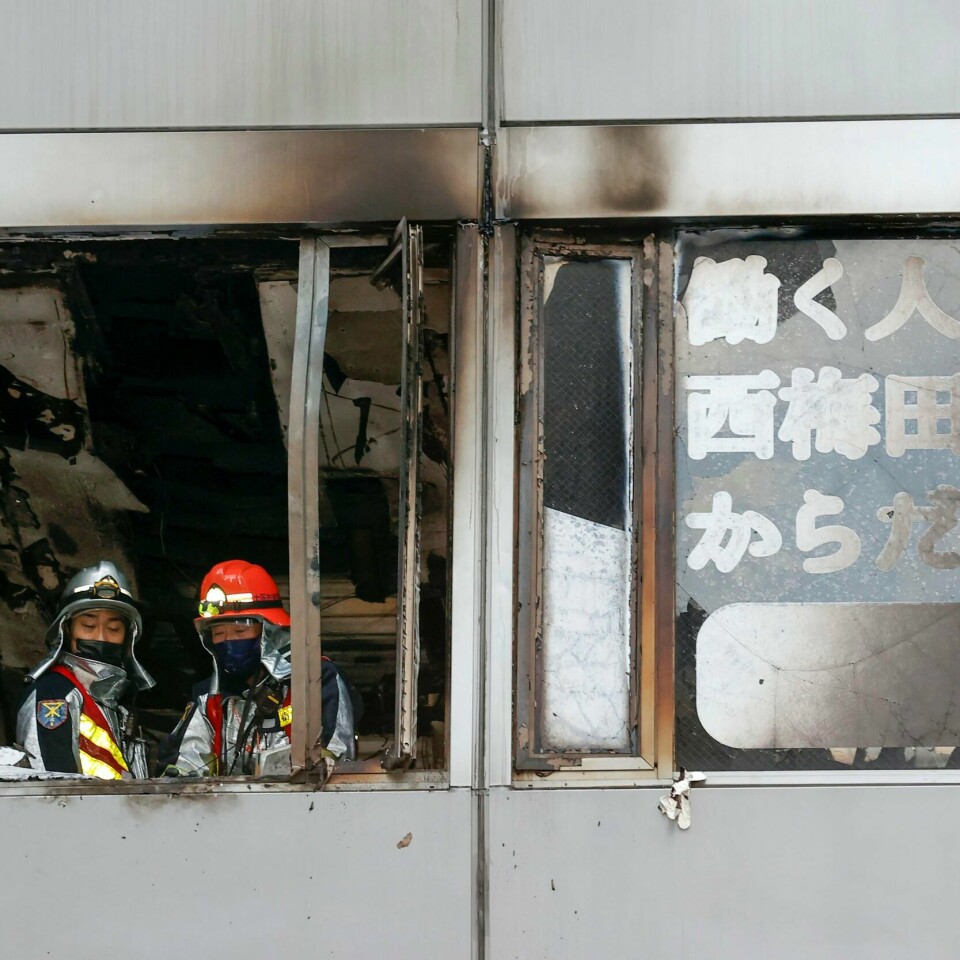 BRANT: Brannmenn står og ser ut av et vindu i bygningen som brant. Brannmenn i vinduet i etasjen der brannen startet. Foto: Kyodo News via AP / NTB