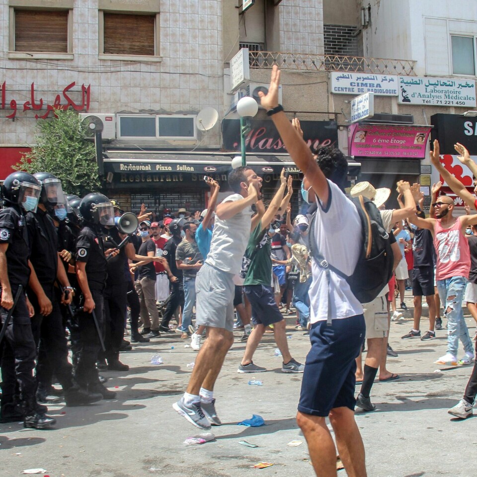 URO: Flere hundre demonstranter samlet seg i Tunisias hovedstad Tunis i helga. De hadde som krav at regjeringen måtte slutte. Søndag sa presidenten at regjeringen var fjernet. Bildet er av folk som protesterer i gatene i Tunis. Det er hovedstaden i Tunisia. De står ansikt til ansikt med politifolk med hjelmer og køller. Foto: Hassene Dridi / AP / NTB