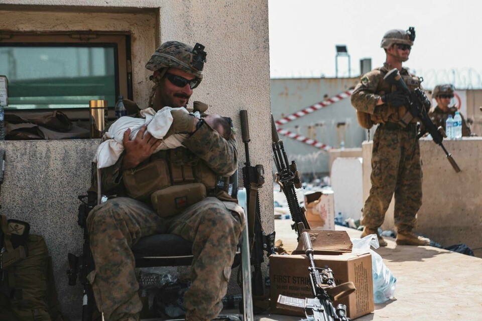 BARN: Danmark vil ikke evakuere barn de ikke vet identiteten til. Bilder viser en amerikansk soldat med en baby i fanget. To menn i ført militært antrekk. En sitter på en stol, mens en står. Militæren som sitter på stolen har en baby på fanget. Han forsøker å roe den ned.