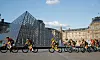 Bildet viser syklister som kjører foran glasspyramiden over plassen foran museet Louvre.