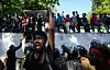 Bildet er av folk som protesterer i Sri Lanka. Det står folk oppe på en mur i bakgrunnen. I front står en mann og roper inn i en megafon mens han holder en hånd i været. Foto: Eranga Jayawardena / AP / NTB