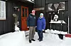 Bildet er av et eldre ektepar som står foran en mørk brun hytte med rød dør. De har på vinterklær og luer. Det er snø på bakken og en drøy halvmeter opp langs veggen på hytta.