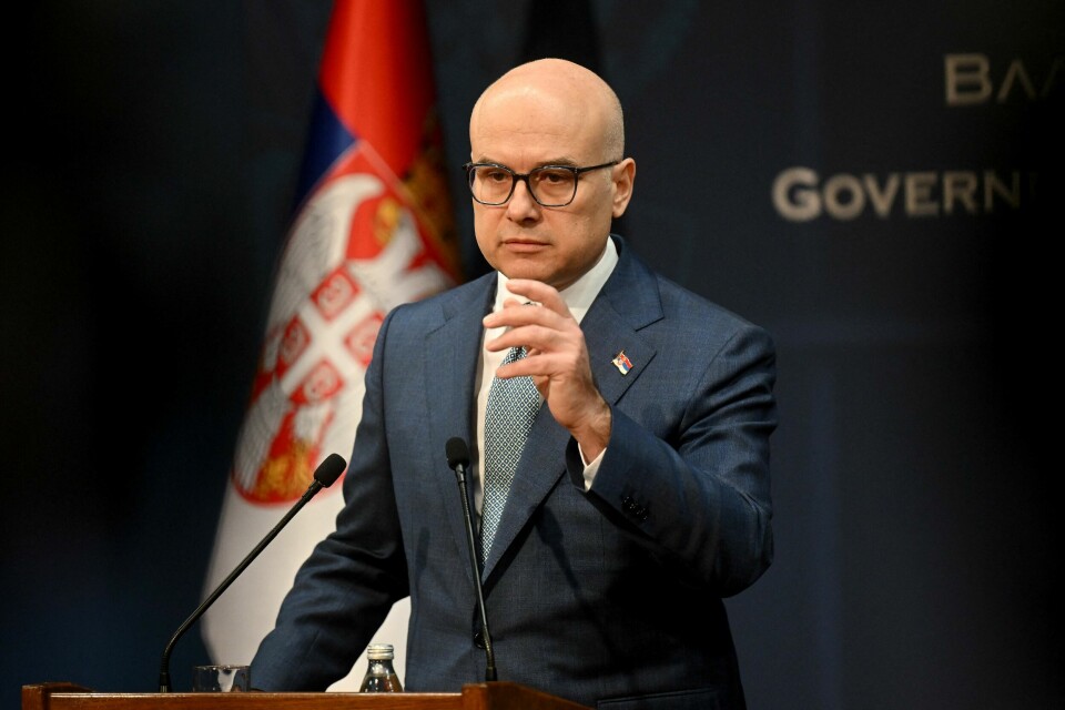 SLUTTER: Serbias statsminister Milos Vucevic slutter. Bildet viser statsministeren til Serbia Milos Vucevic i dress og blått slips. Bak ham er det serbiske flagget.