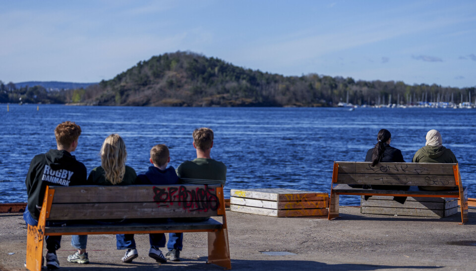 SOL: Mange nyter vårsola ved sjøen. Denne uka blir det mildt og fint vær i hele landet. Seks personer sitter på benker og ser ut over havet på en solrik dag. Det er blå himmel, klart vann og øyer i bakgrunnen.