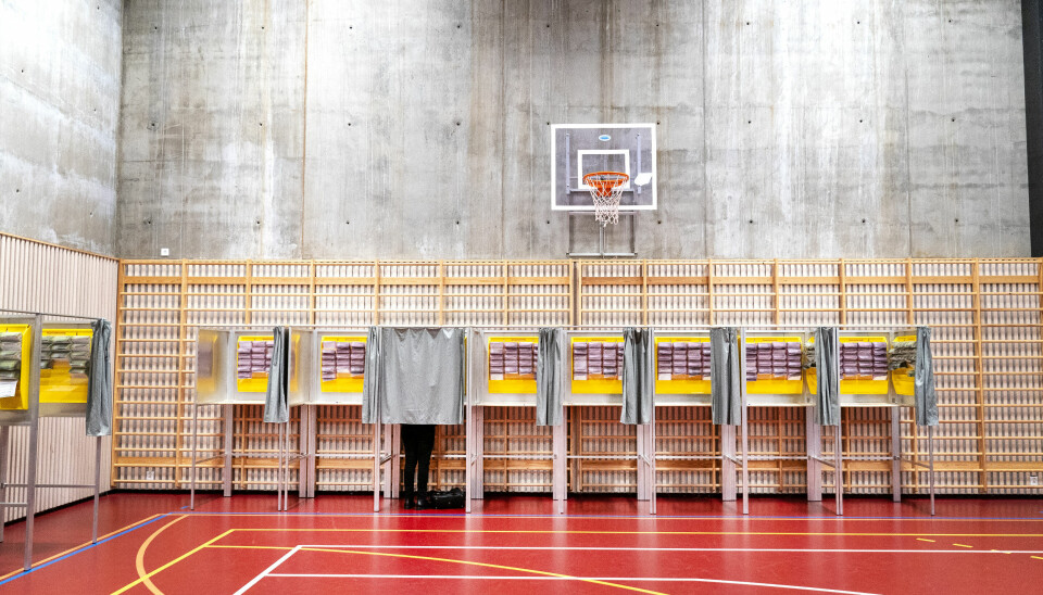 STEMME: Ved høstens valg avgjør vi hvem som skal styre Norge. Valglokale i en gymsal med flere stemmebokser på rad. Én person står inne i en av boksene bak et forheng. Over boksene henger en basketballkurv på en betongvegg.