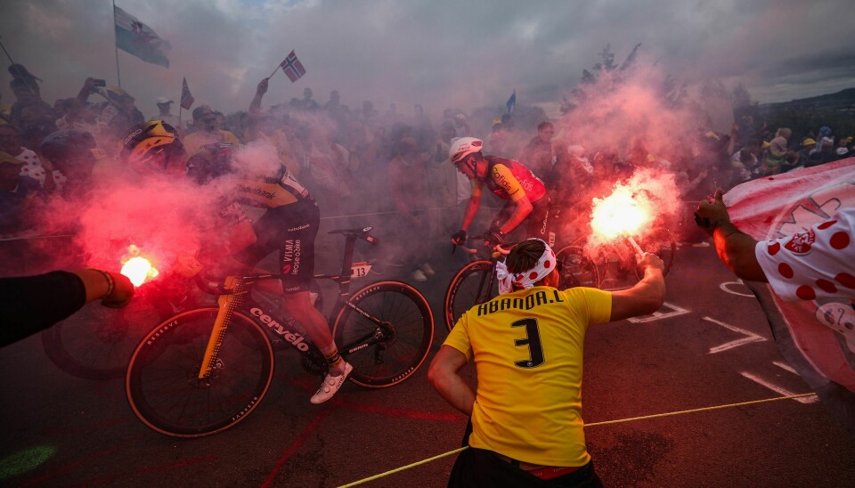 FOLKEFEST: Tour de France er en folkefest. Syklistene må ofte presse seg gjennom folkemengder som tar litt for mye plass. Touren går fra 5. juli til 27. juli. Syklister kjemper seg opp en bratt bakke under Tour de France. Rundt dem er det folk som holder opp bluss med rød røyk.