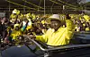 President Yoweri Museveni i gul skjorte og hatt gir tommel opp til folket fra en bil. Alle rundt har også på gul skjorte eller t-skjorte.
