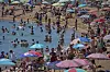 Mange folk bader og soler seg på en stappfull strand i Spania. Det er tett med mennesker både i vannet og på sanden. Flere bruker fargerike parasoller for å skjerme seg mot sola.