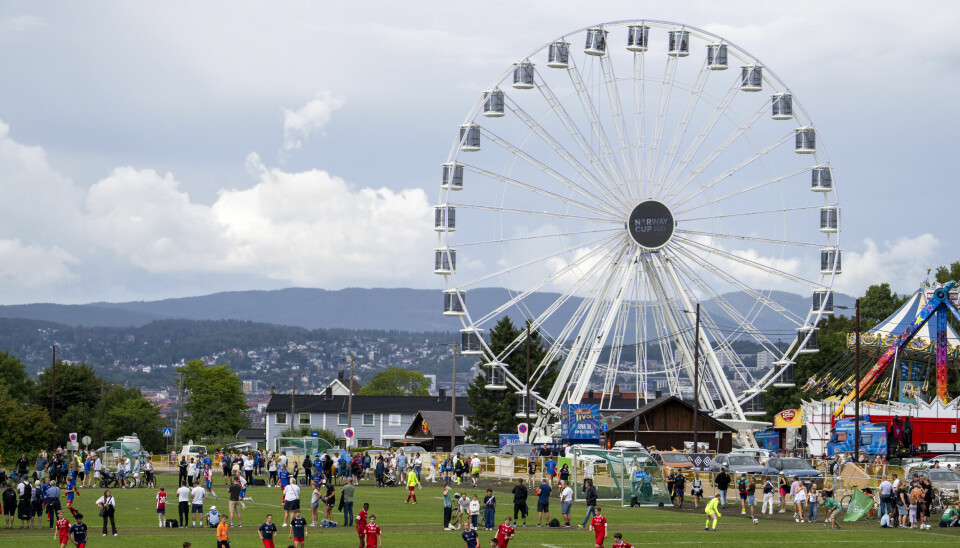 FULL FART: Norway Cup byt både på fotball og moro. Fotballspillere og publikum på en gressbane under Norway Cup, med et stort pariserhjul og tivoli i bakgrunnen.