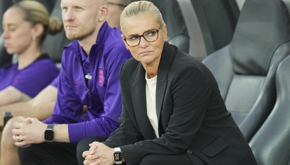 FOTBALL: Sarina Wiegman har ledet England til tre finaler på rad. Søndag kan hun vinne sitt tredje EM-gull som trener. Sarina Wiegman sitter på benken med et alvorlig blikk, kledd i en svart dressjakke og briller.